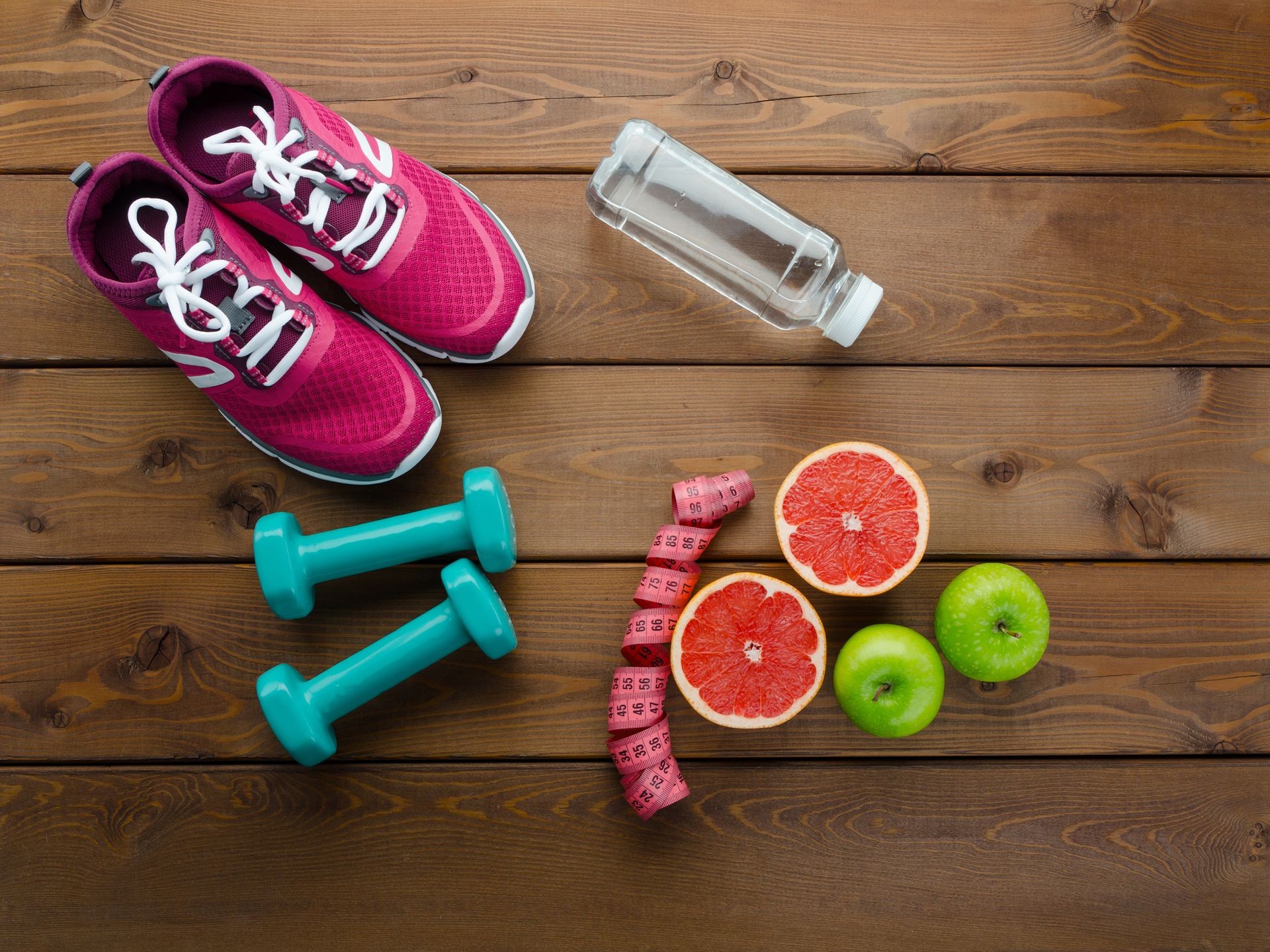 Fitness concept with sneakers dumbbells bottle of water apple and measure tape on wooden table background  